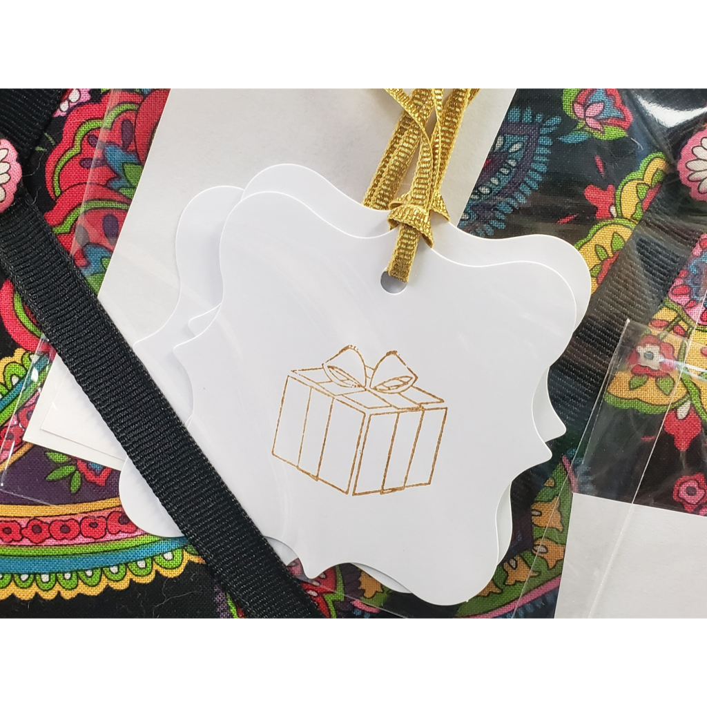 A handmade white gift tag with a picture of a gold stamped gift box with gold ribbon for attaching to gifts.  Located in Gem Designs' Booth inside Antiques & Artisans at 619 Trolley Rd, Summerville in the Christmas tree room.