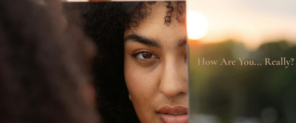 Woman looking at her reflection in a mirror at sunrise with the words "How Are You... Really?" encouraging a quiet moment of self-care and personal reflection.