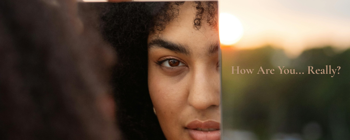 Woman looking at her reflection in a mirror at sunrise with the words "How Are You... Really?" encouraging a quiet moment of self-care and personal reflection.